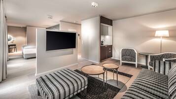 A modern hotel room with a large bed, a sofa, a TV, a small kitchen area, and a seating area with a lamp.