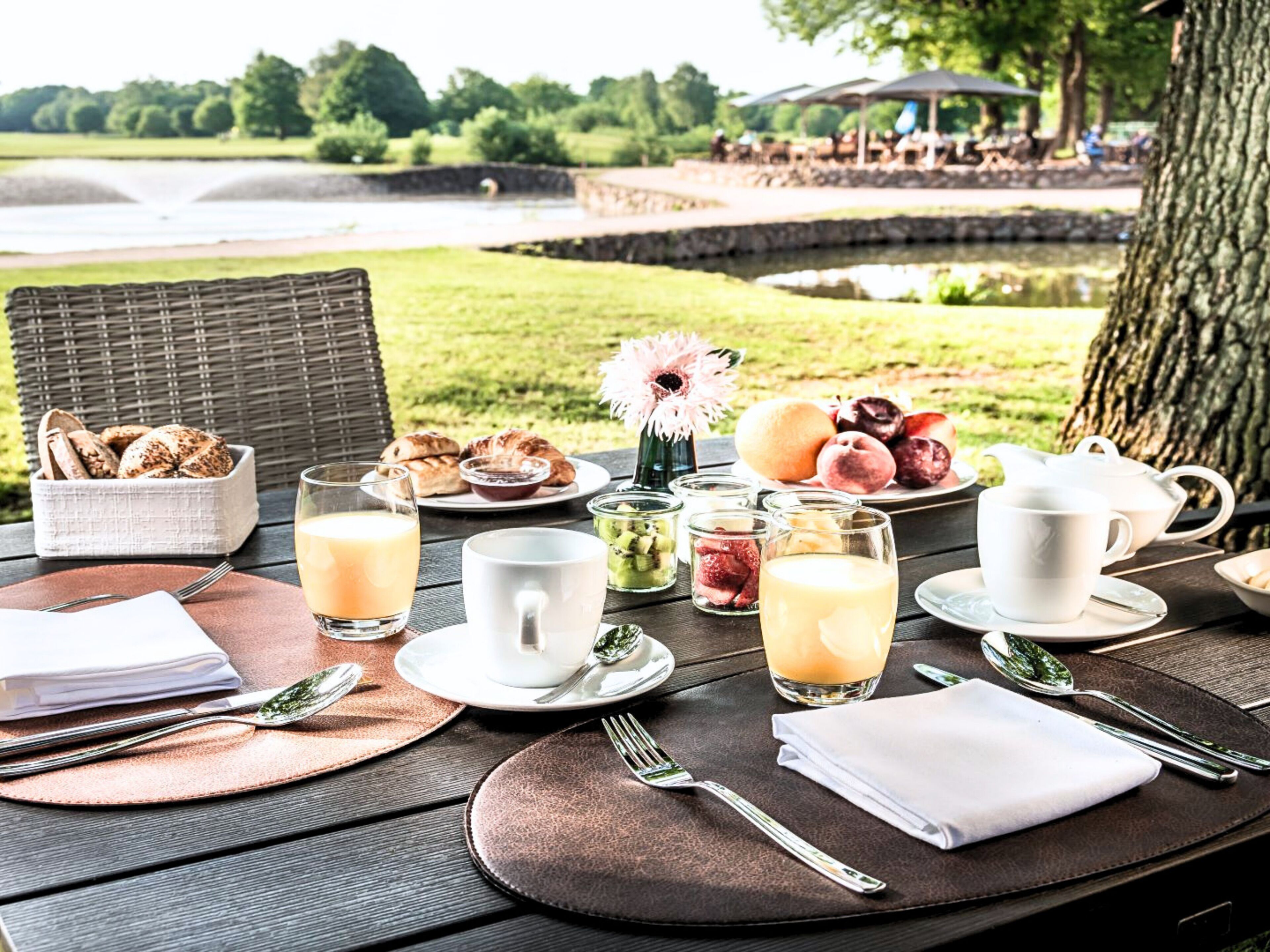 Amanecer con vistas a un lago y árboles, con pan, bollería, frutas y bebidas sobre una mesa de madera.