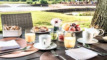 Amanecer con vistas a un lago y árboles, con pan, bollería, frutas y bebidas sobre una mesa de madera.