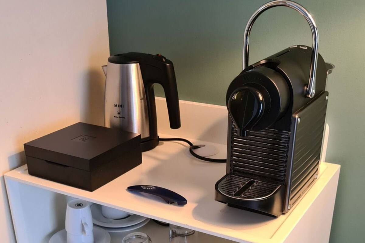 A kitchen area with a coffee maker, a kettle, a box, and a remote control on a white shelf against a teal wall.
