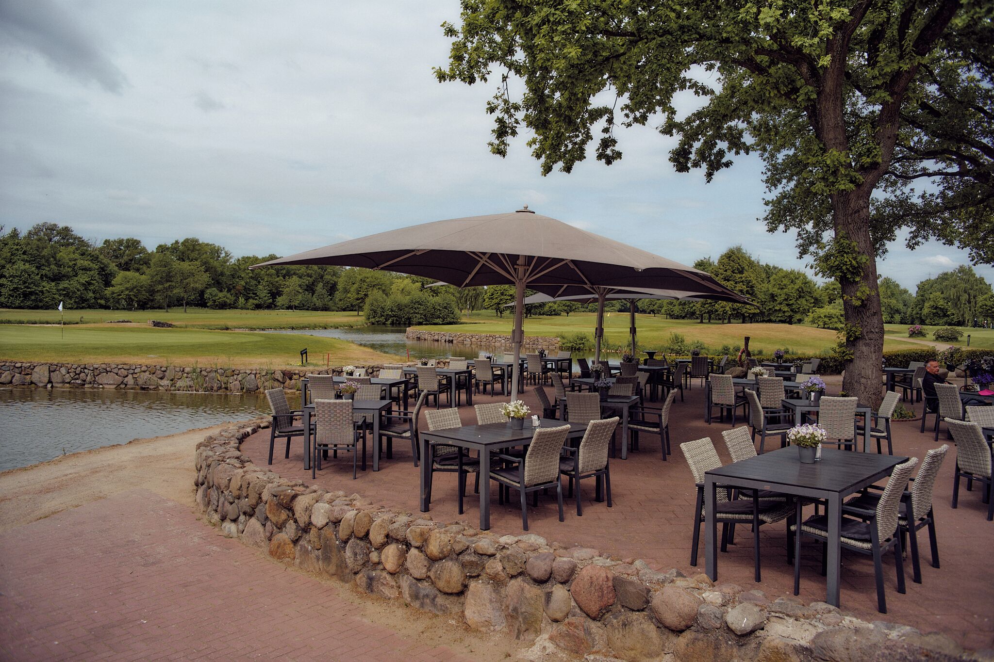 An outdoor dining area with tables and chairs, a large umbrella, and a view of a golf course and pond.