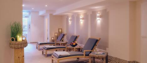 Un espacio de spa con múltiples sillones de relax, una mesita de madera con velas y una silla de mimbre con bandeja.