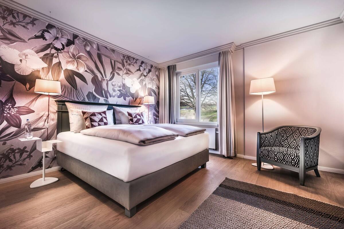 A modern bedroom with a large bed, a nightstand, a chair, and a window with curtains.