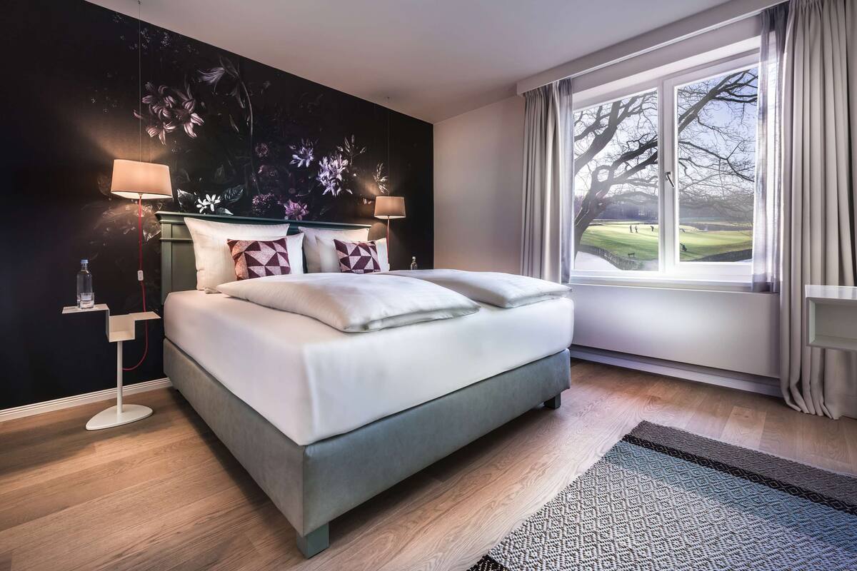 Un dormitorio moderno con cama, dos lámparas de noche, una mesita de noche, un ventanal con cortinas y un mural en la pared.