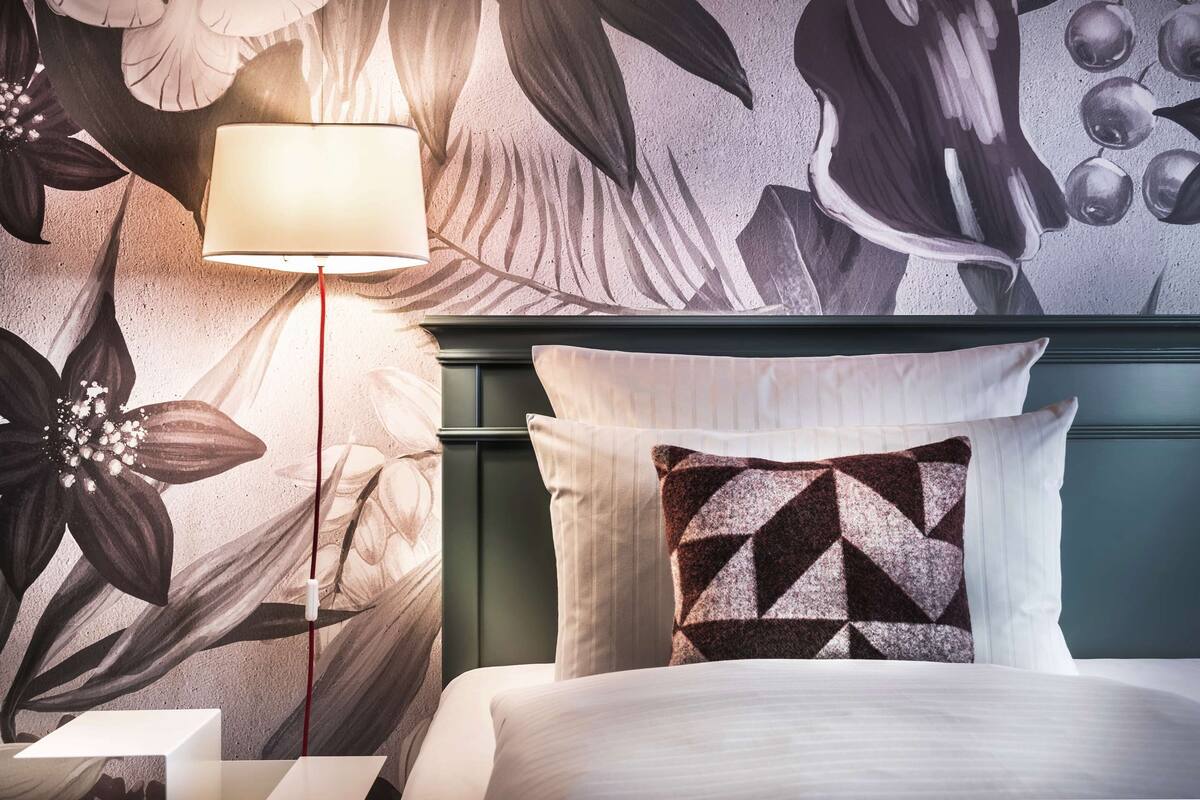 Habitación de hotel con un mural floral en la pared, una cama con ropa blanca, un cojín de estampado geométrico y una lámpara de pie.