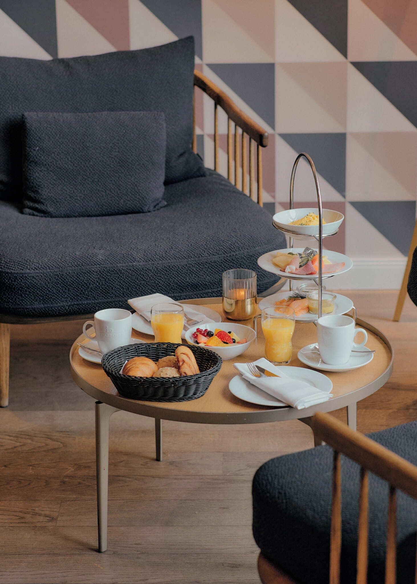 A breakfast setup with a three-tiered serving tray, a basket of pastries, and a coffee table in a modern room with a geometric-patterned wall and a sofa.