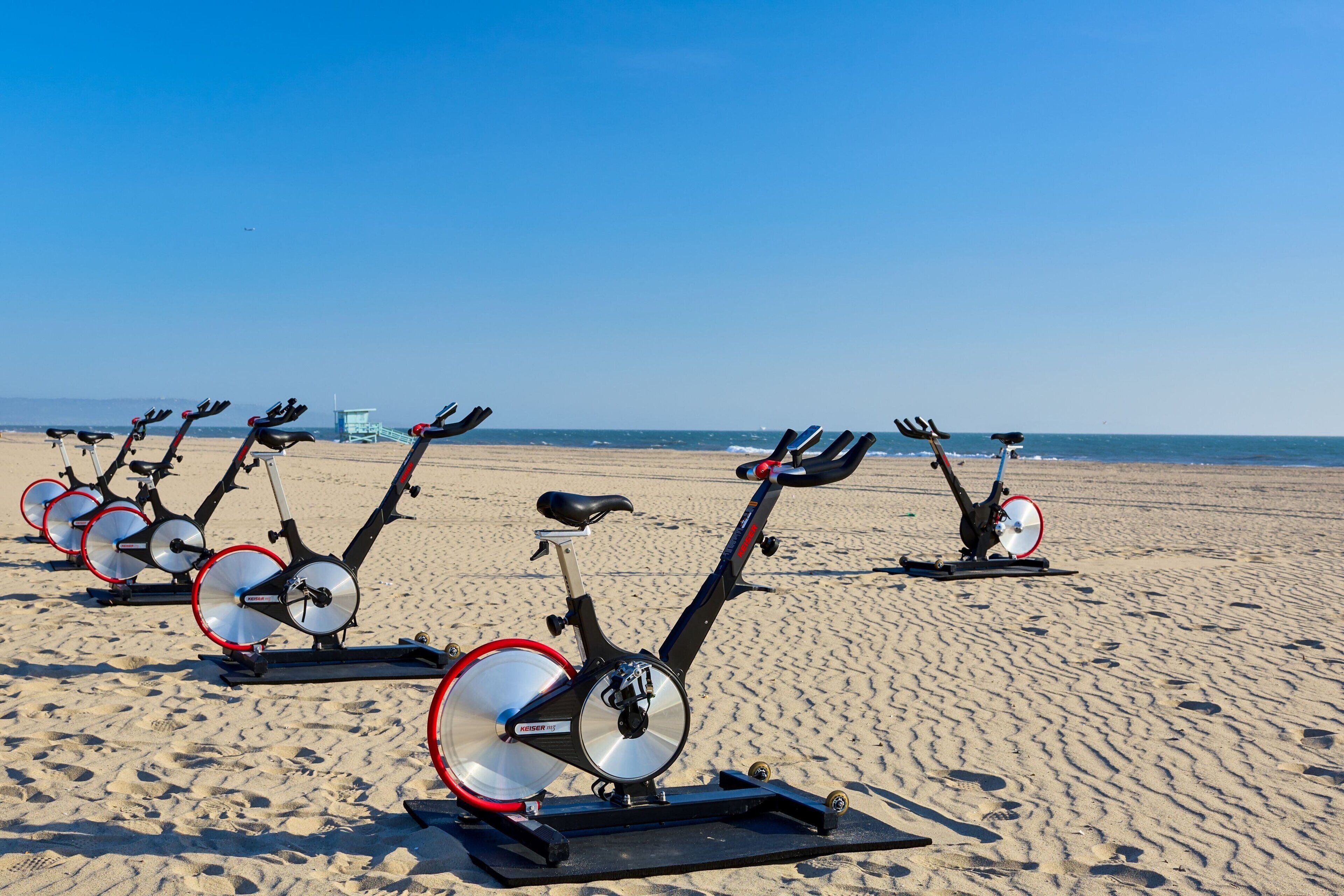 Stationary bikes lined up on a sandy beach with a clear sky in the background.