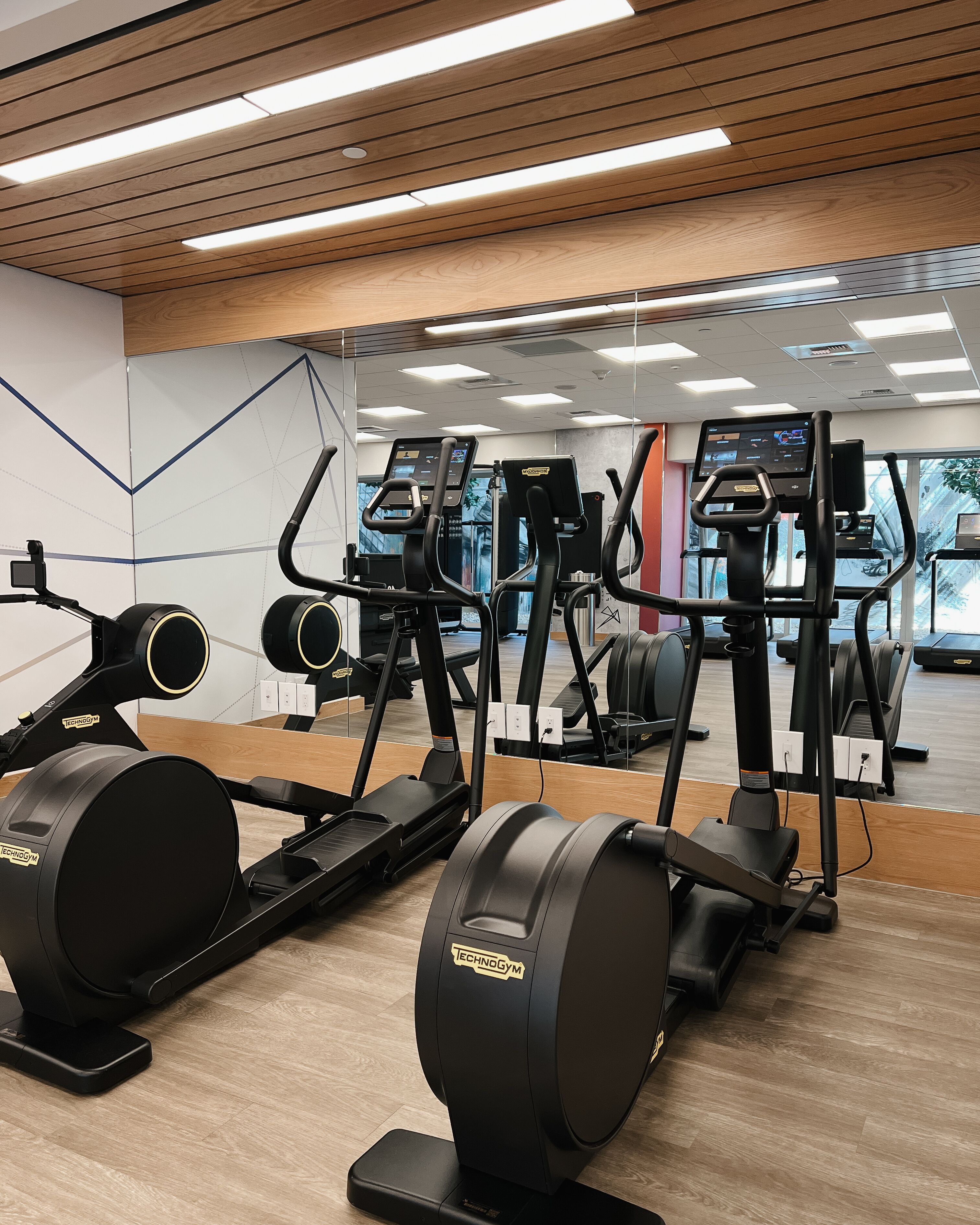 Une salle de sport dotée de nombreux appareils de cardio, notamment des elliptiques et des tapis de course, disposés dans un espace vaste, avec un plancher en bois et de grands miroirs sur les murs.