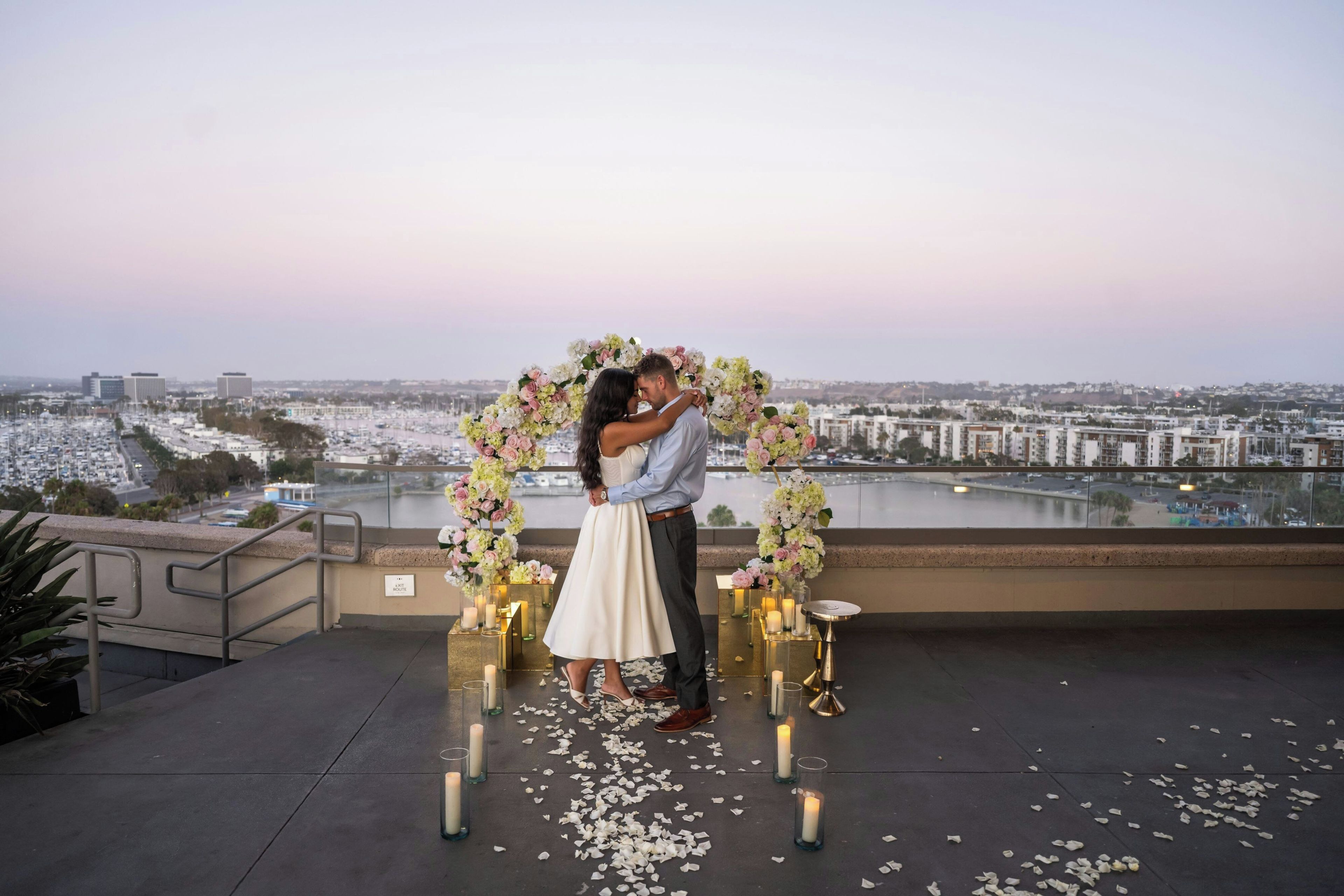 A couple embracing under a floral arch on a rooftop with a city view.