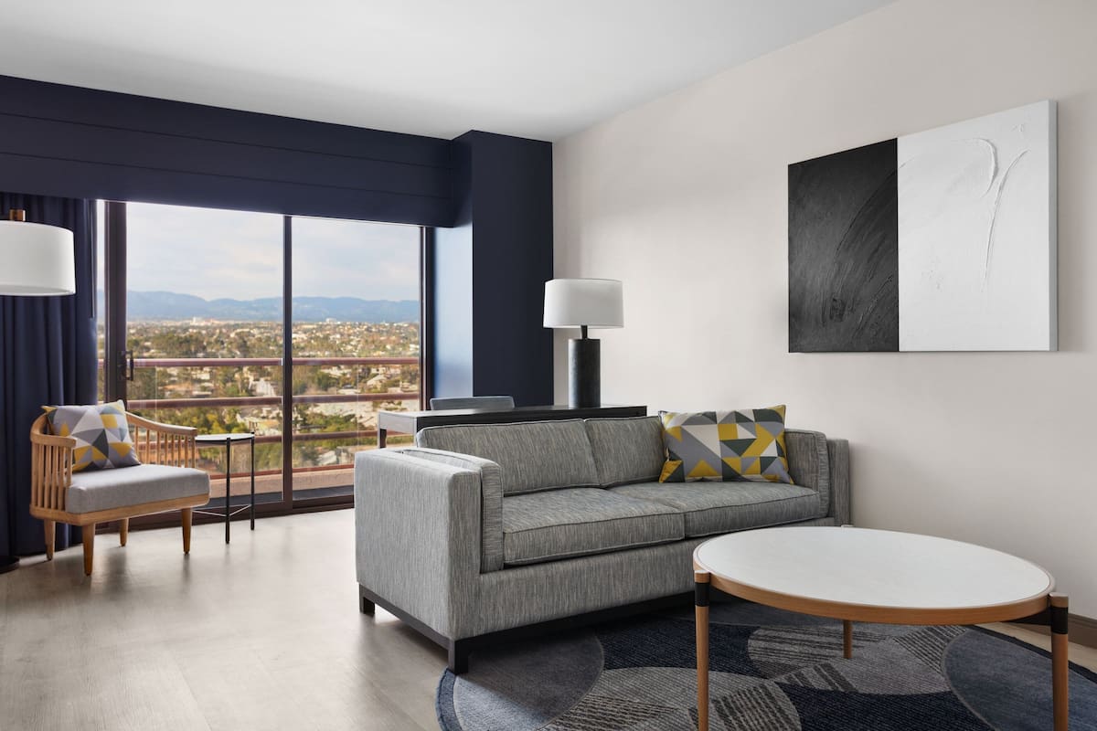 Habitación de hotel moderna con un sofá gris, una mesa redonda y vistas de la ciudad a través de amplias ventanas.