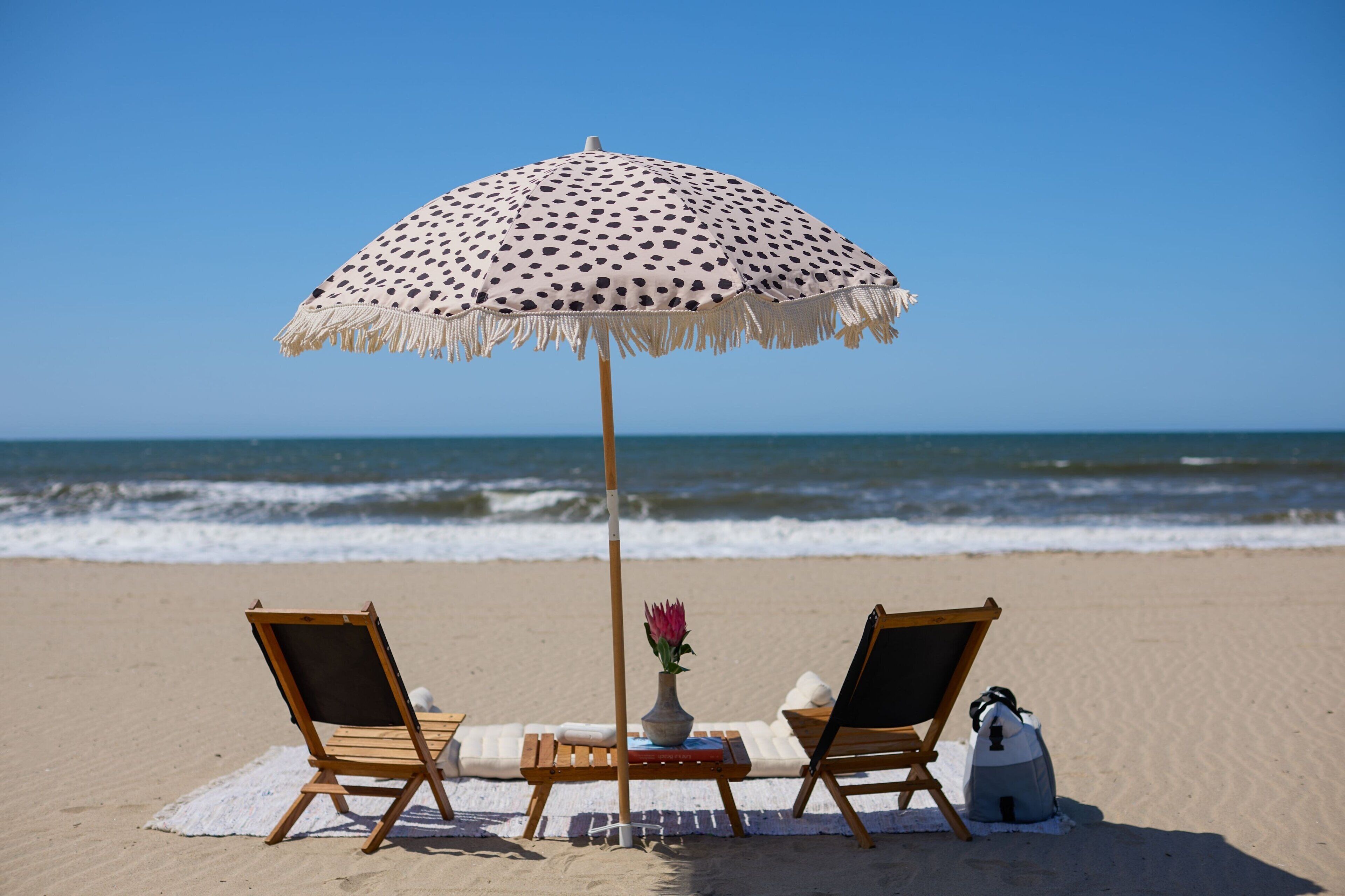 A beach scene with a polka-dotted umbrella, two wooden lounge chairs, a small table with a vase and a book, and a cooler on the sand.