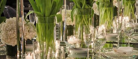 Ett långt bord dukat med kristallglas, vita servetter och ljus, omgivet av blomsterarrangemang och grönska.