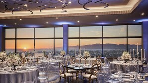 Uma sala de banquetes com mesas redondas, cadeiras transparentes e vista para o pôr do sol através de grandes janelas.