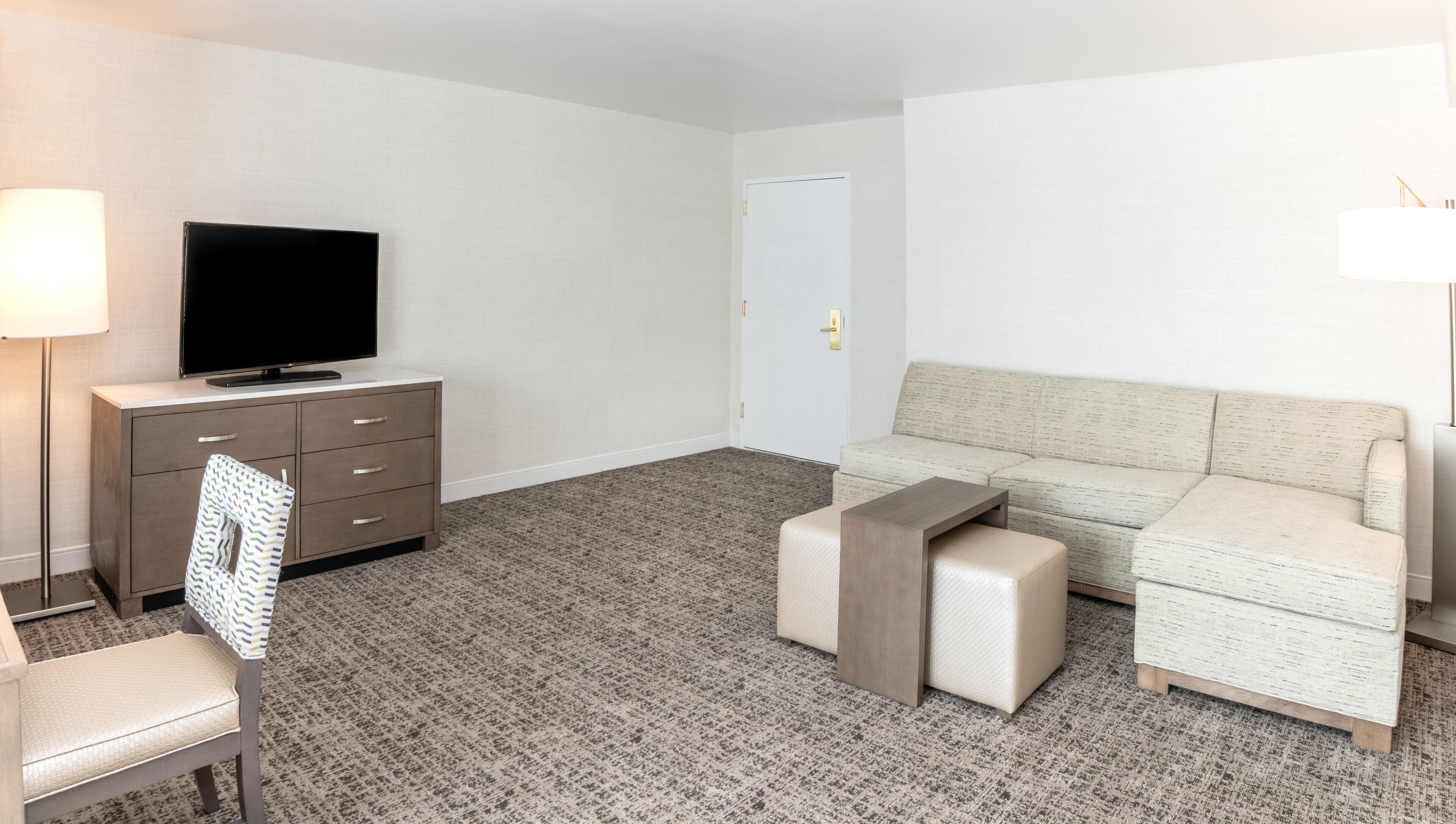 A modern hotel room with a flat-screen TV on a dresser, a sofa, a small ottoman, and a lamp.