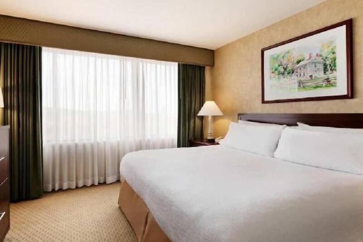 A hotel room with a large bed, a nightstand with a lamp, a painting on the wall, and a window with curtains.