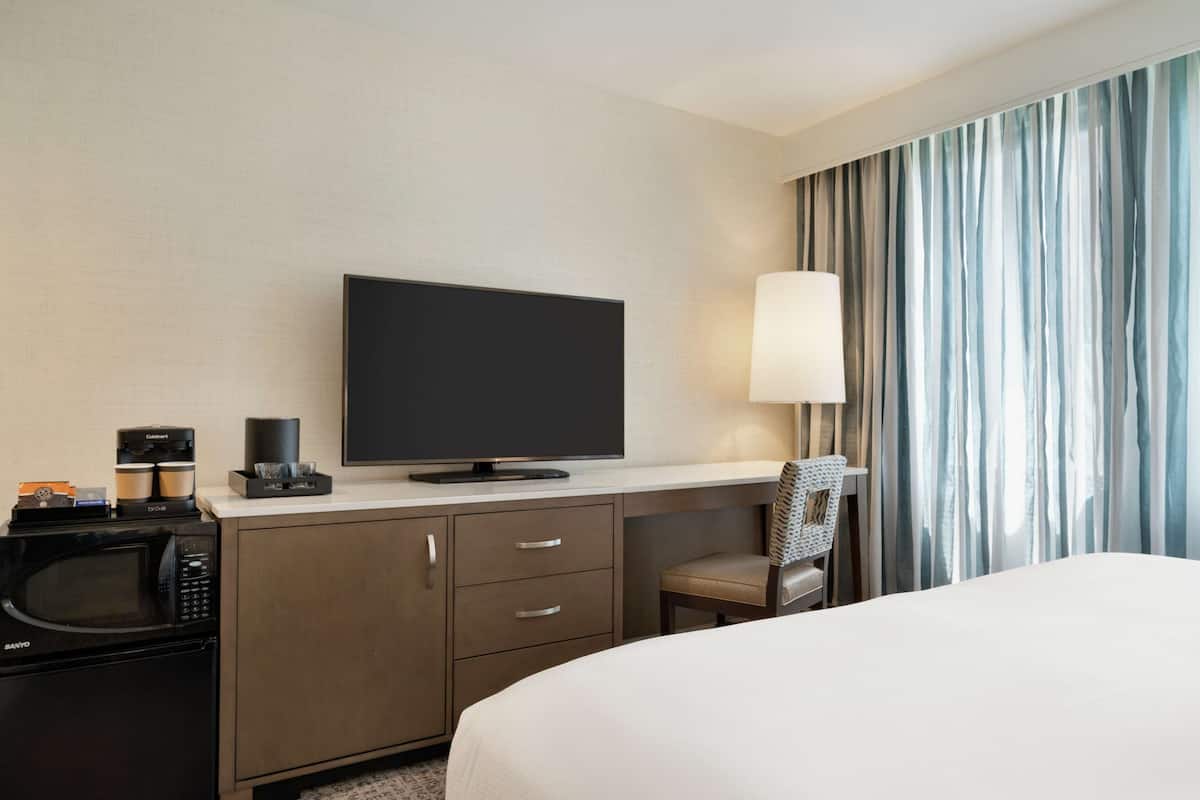 A hotel room with a bed, a TV, a coffee maker, a microwave, a desk with drawers, a chair, and a window with curtains.