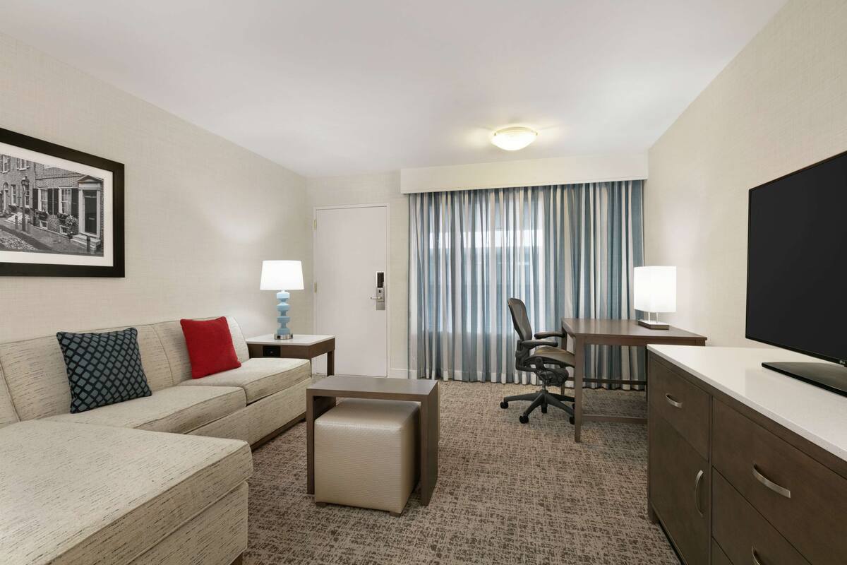 A hotel room with a sofa, a desk, a chair, a TV, a lamp, and a framed picture on the wall.