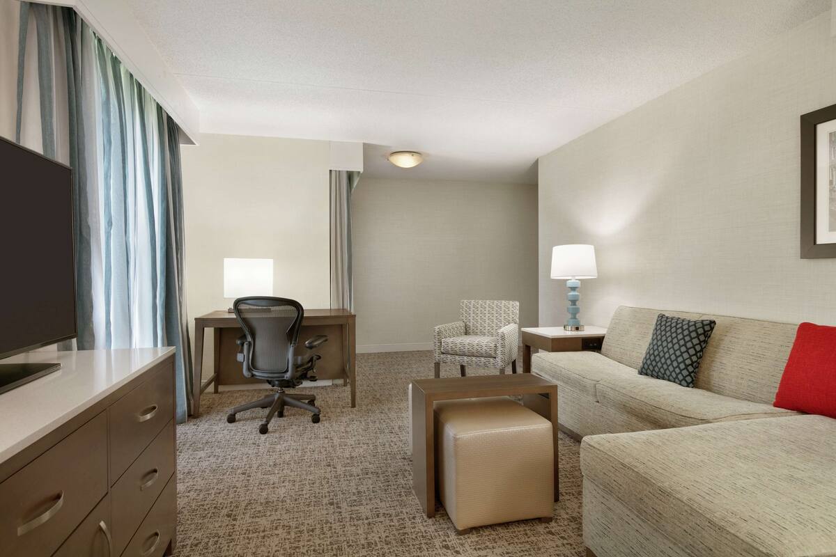 A hotel room with a desk, office chair, sofa, ottoman, and a television.