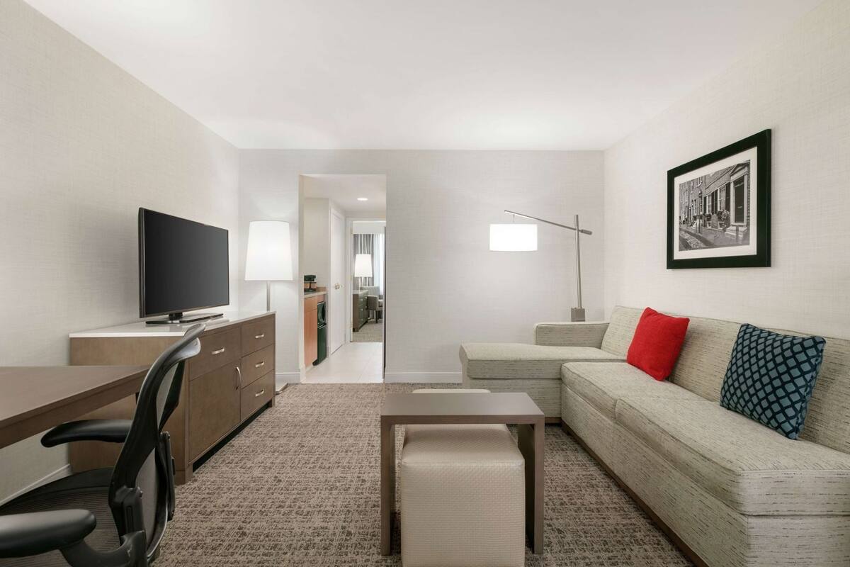 A modern living room with a flat-screen TV, a sofa with patterned cushions, a desk with a chair, and a floor lamp.