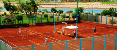Un court de tennis avec un filet, entouré d’une clôture, et offrant une vue sur une étendue d’eau et des montagnes en arrière-plan.