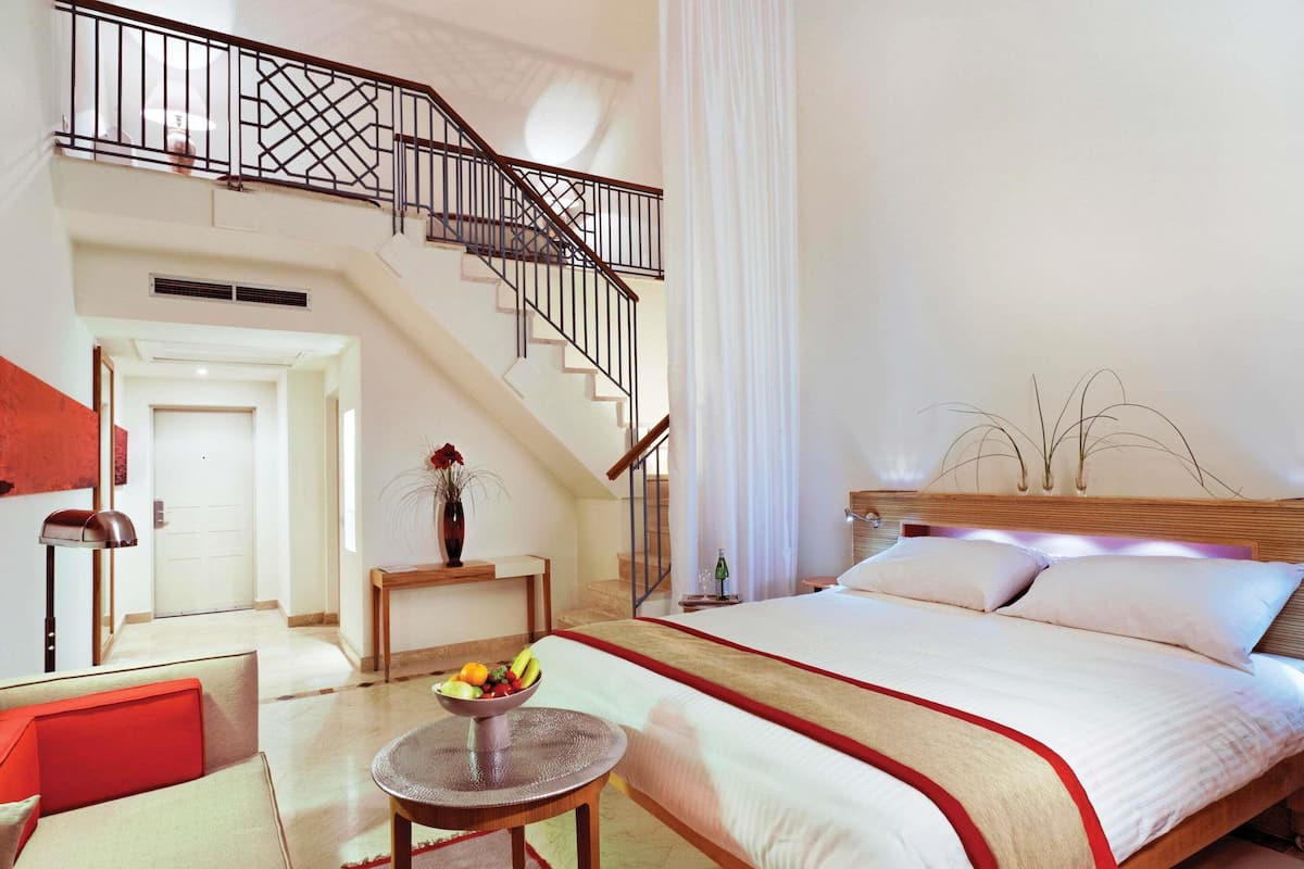 A bedroom with a bed, a small table with a bowl of fruit, a nightstand, a lamp, a staircase, and a door.