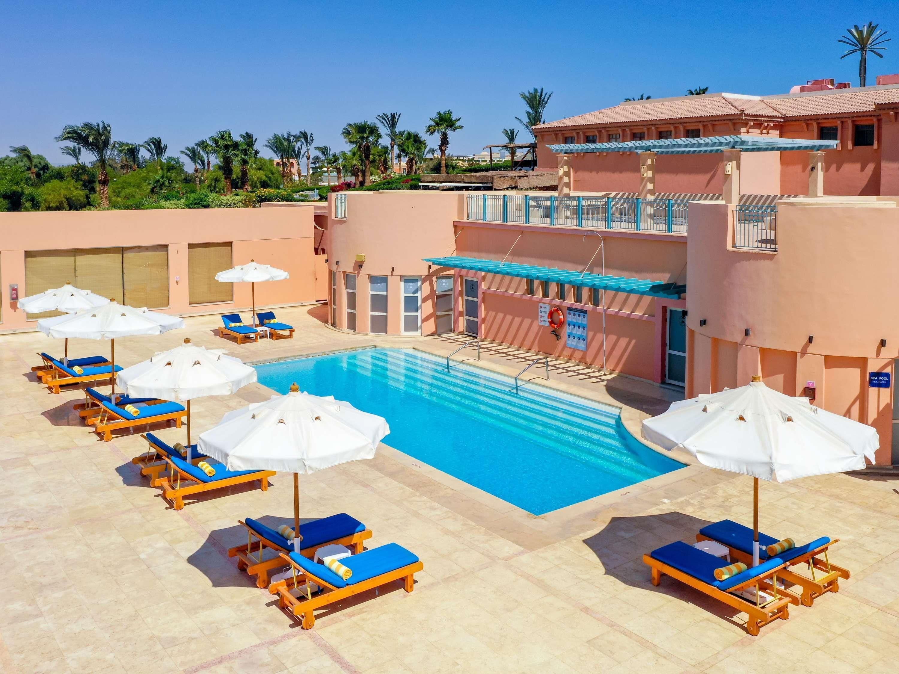 A pool area with multiple sun loungers and umbrellas, a clear blue pool, and a building with a tiled roof.