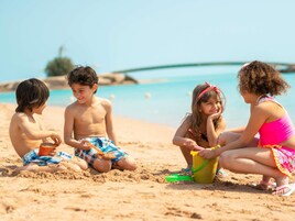 Niños jugando en una playa arenosa con un cielo azul despejado y un mar tranquilo al fondo.