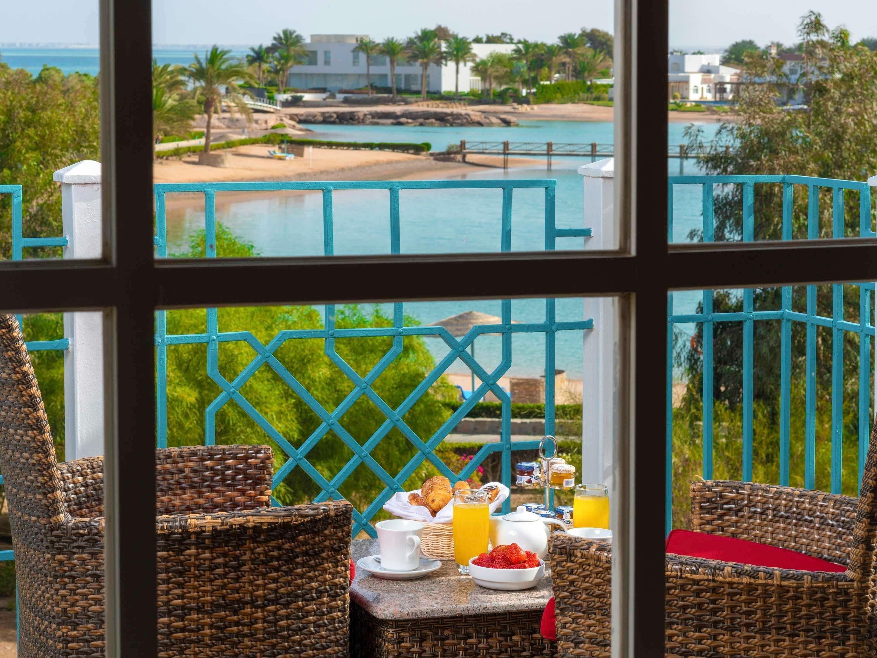 A balcony with wicker chairs, a table with breakfast items, and a view of a beach and pool.