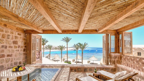 Une pièce avec un mur en pierre, un plafond en bois, une table en verre, une chaise en bois, un lit en bois et une vue sur la plage et l’océan.