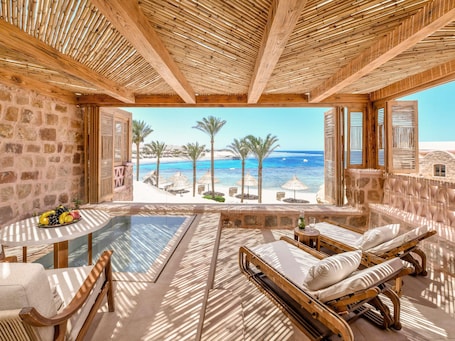A room with a stone wall, wooden ceiling, a glass table, a wooden chair, a wooden bed, and a view of the beach and ocean.