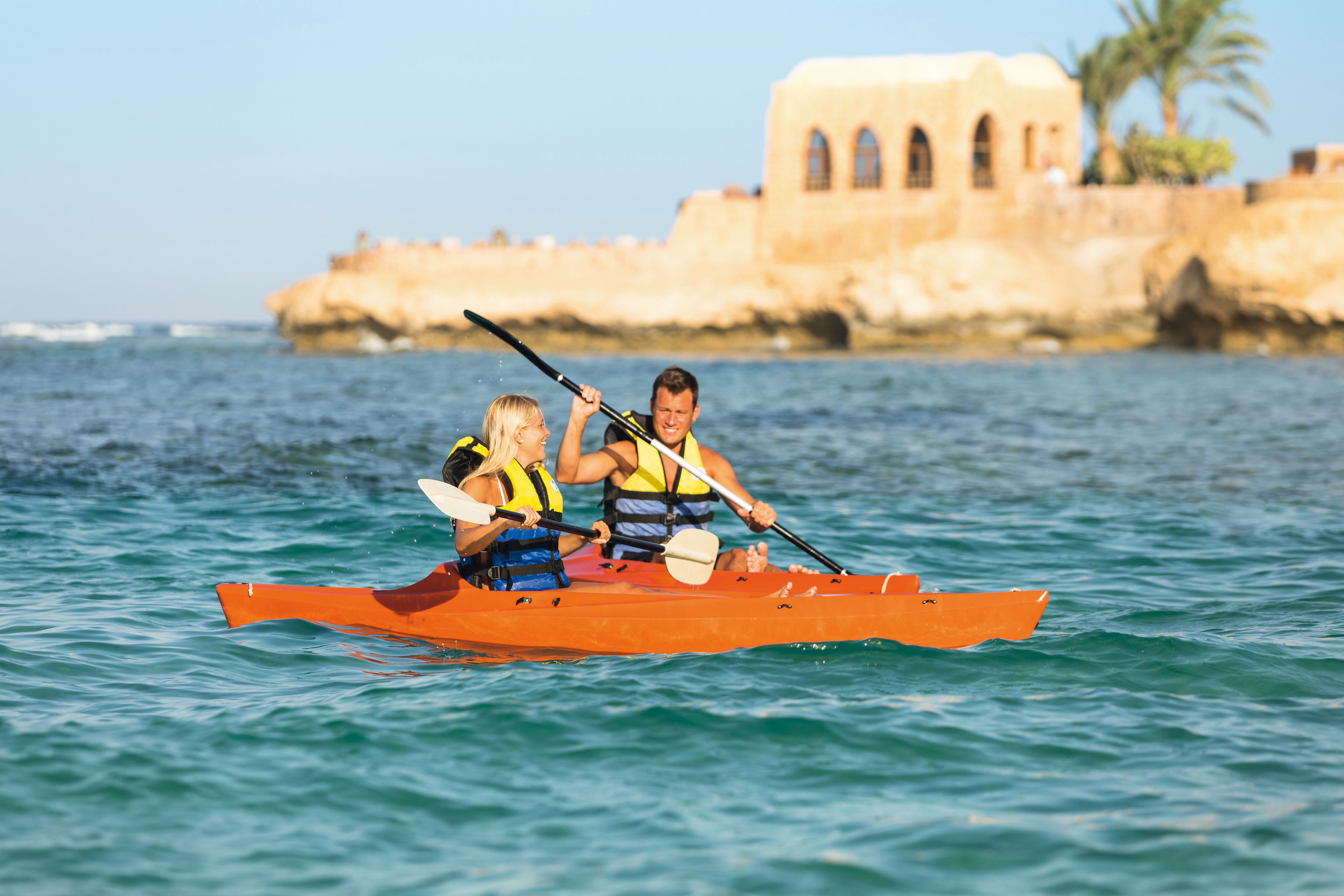 Ein Paar sitzt in einem kleinen, orangefarbenen Kajak auf ruhigem Wasser, im Hintergrund ist ein Küstenhaus zu sehen.