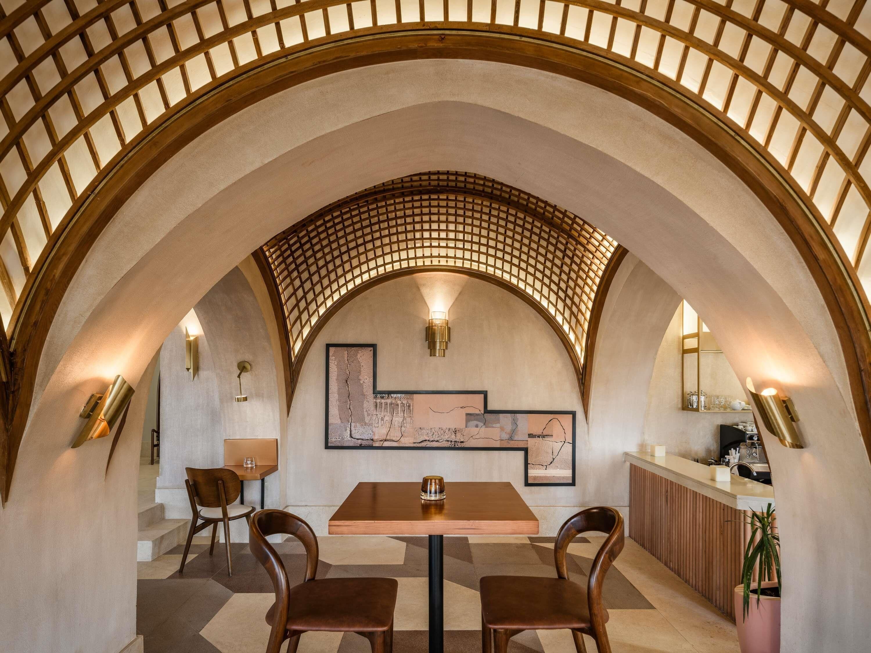Een gewelfde plafond met een rasterpatroon, een tafel met twee stoelen, een schilderij aan de muur, en een bar met een toonbank en krukken.