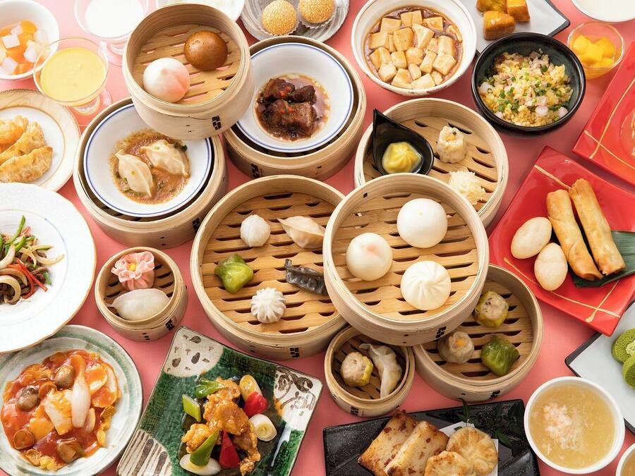 Diversos pratos asiáticos, incluindo dumplings, tofu e uma tigela de sopa, dispostos sobre uma mesa rosa.
