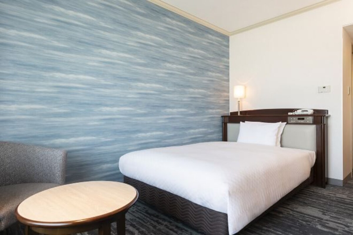 Habitación de hotel con una cama, una silla, una mesita, una mesita de noche y una pared con diseño texturizado.