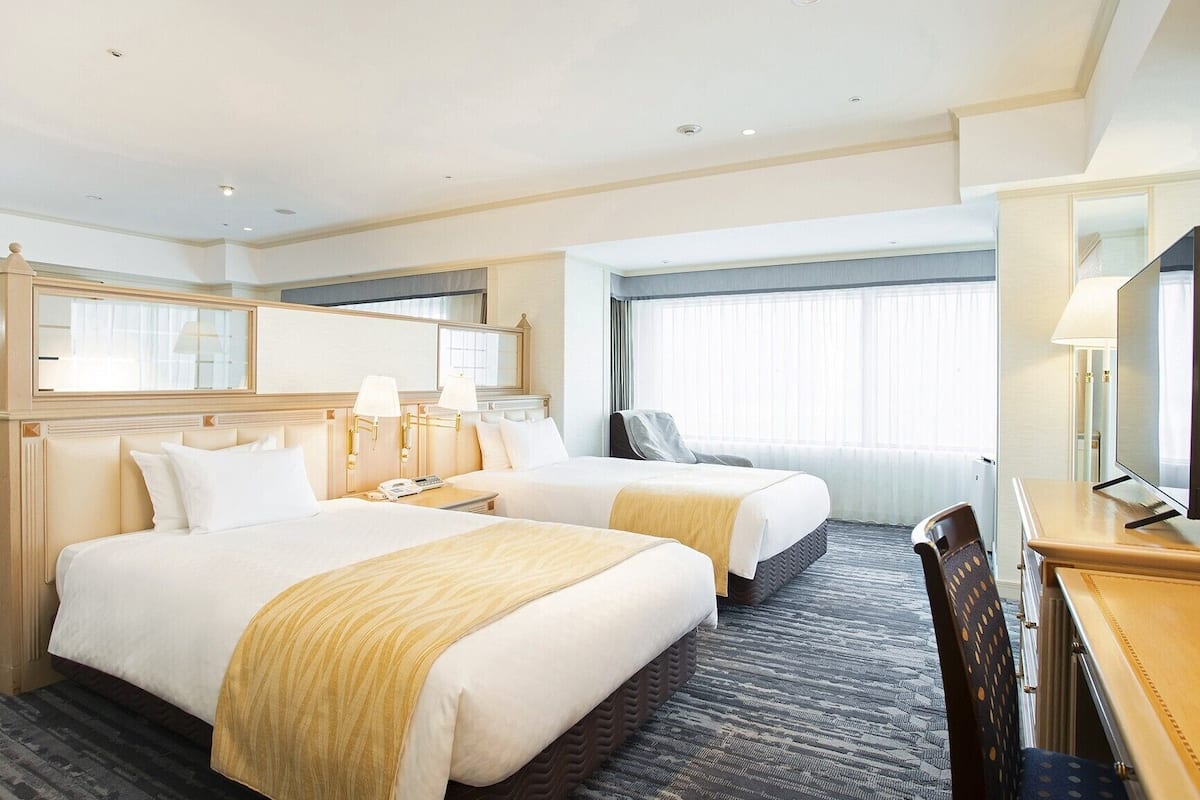 A hotel room with two beds, a desk, a chair, a TV, and a window with curtains.