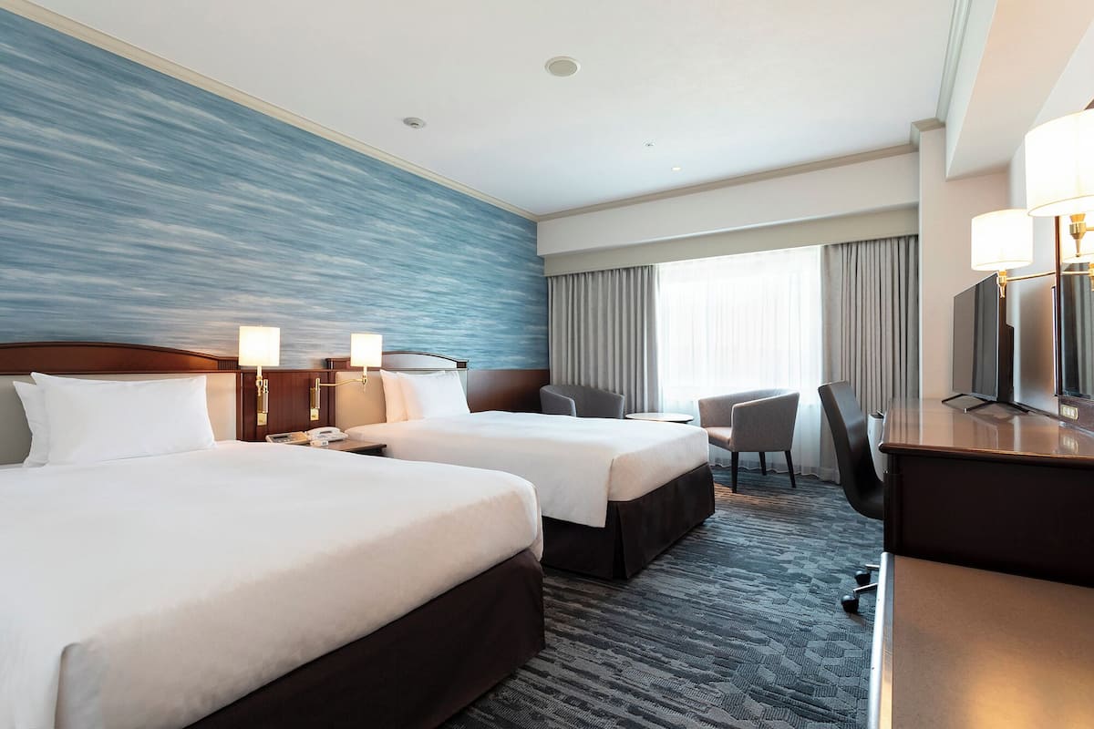 A hotel room with two beds, a desk, a chair, a TV, and a large window with curtains.