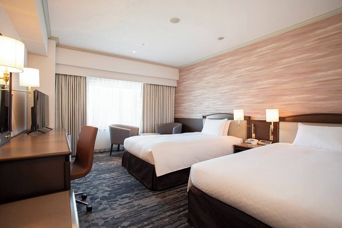 A hotel room with two beds, a desk, a chair, and a television.