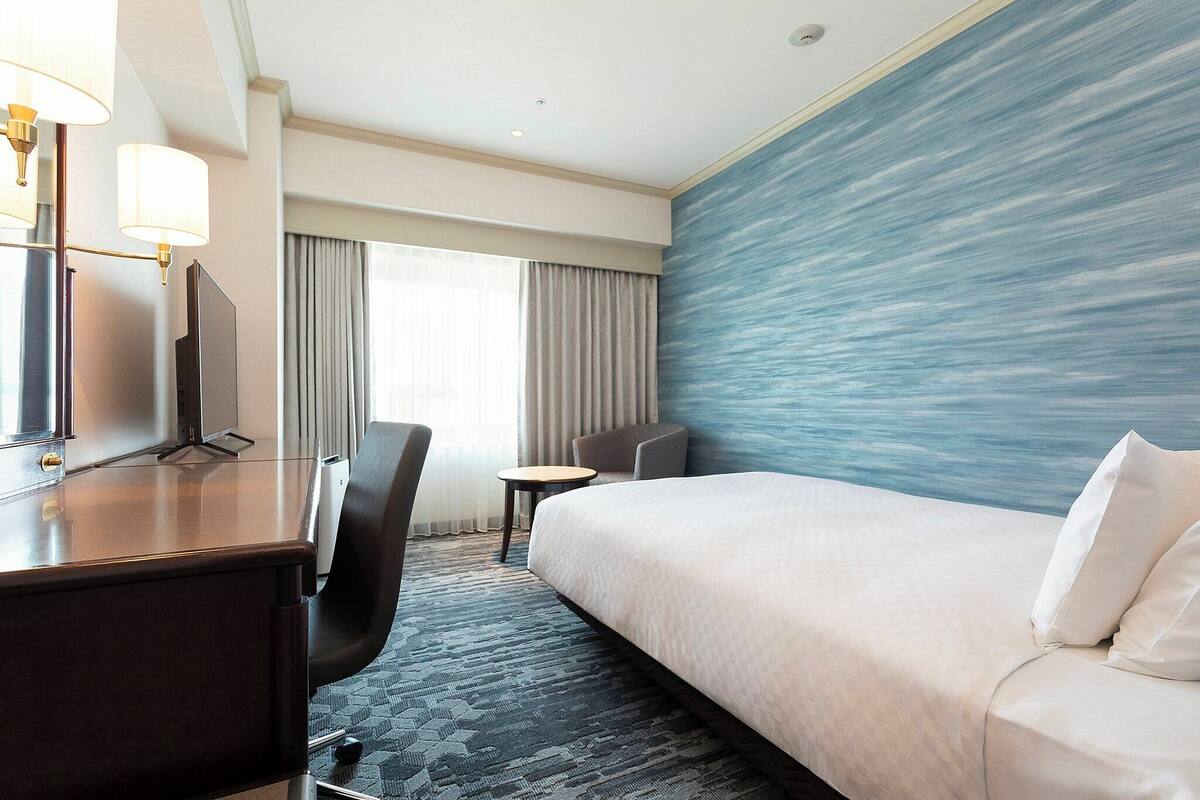 A hotel room with a bed, a desk with a computer, a chair, a small table, and a view of the sea through the window.