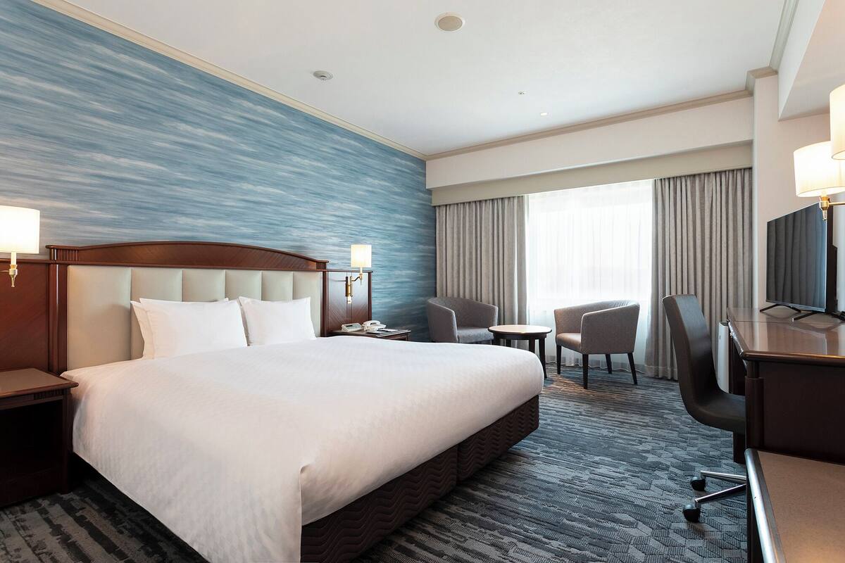 A hotel room with a large bed, a desk with a chair, a small table, and a TV.