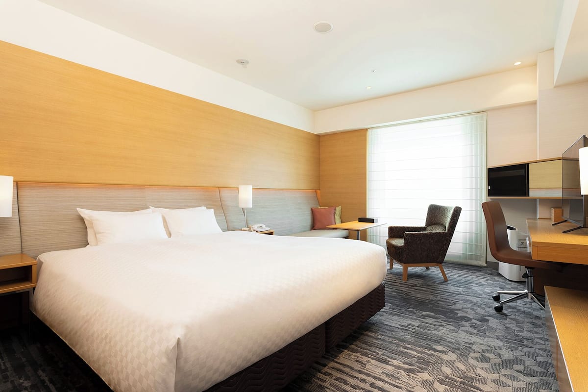 A hotel room with a large bed, a desk with a chair, a TV, and a window with blinds.