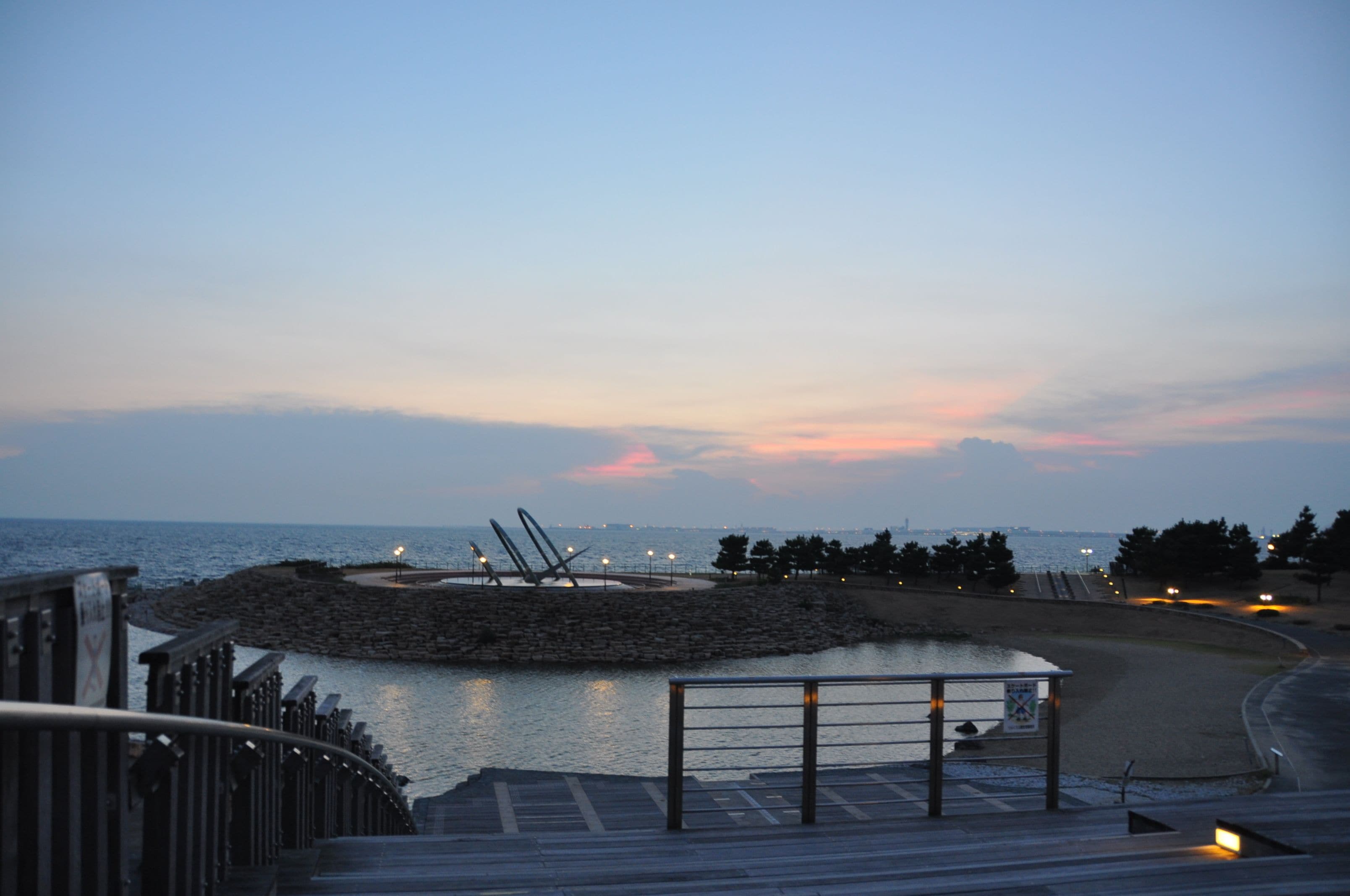 A waterfront promenade with a circular viewing platform, a metal fence, and a paved walkway.