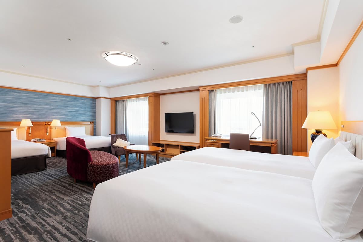 A hotel room with two beds, a desk, a TV, and a seating area with a chair and ottoman.