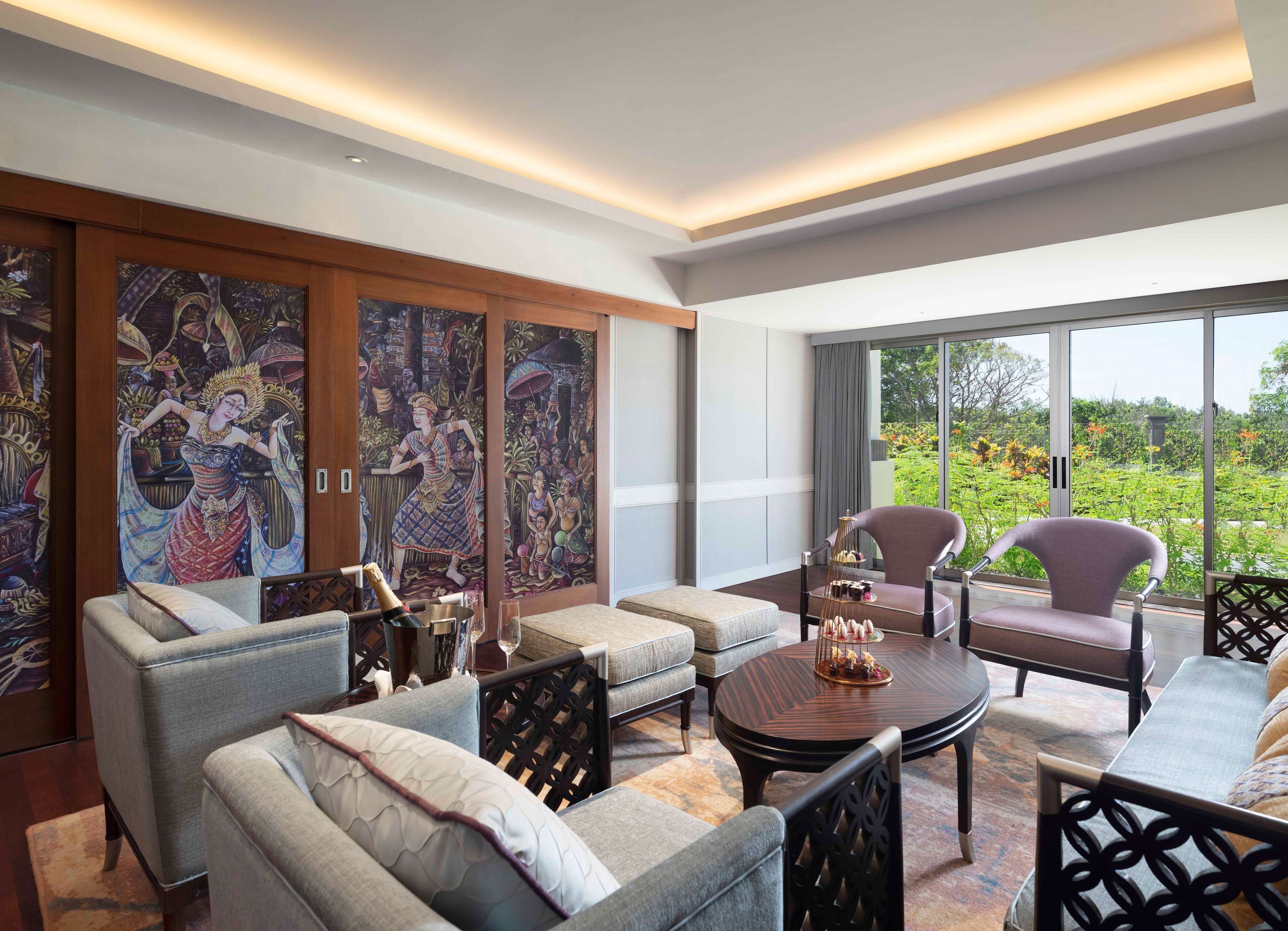 A modern living room with a sofa, armchairs, and a coffee table. There are large windows with a view of greenery outside.