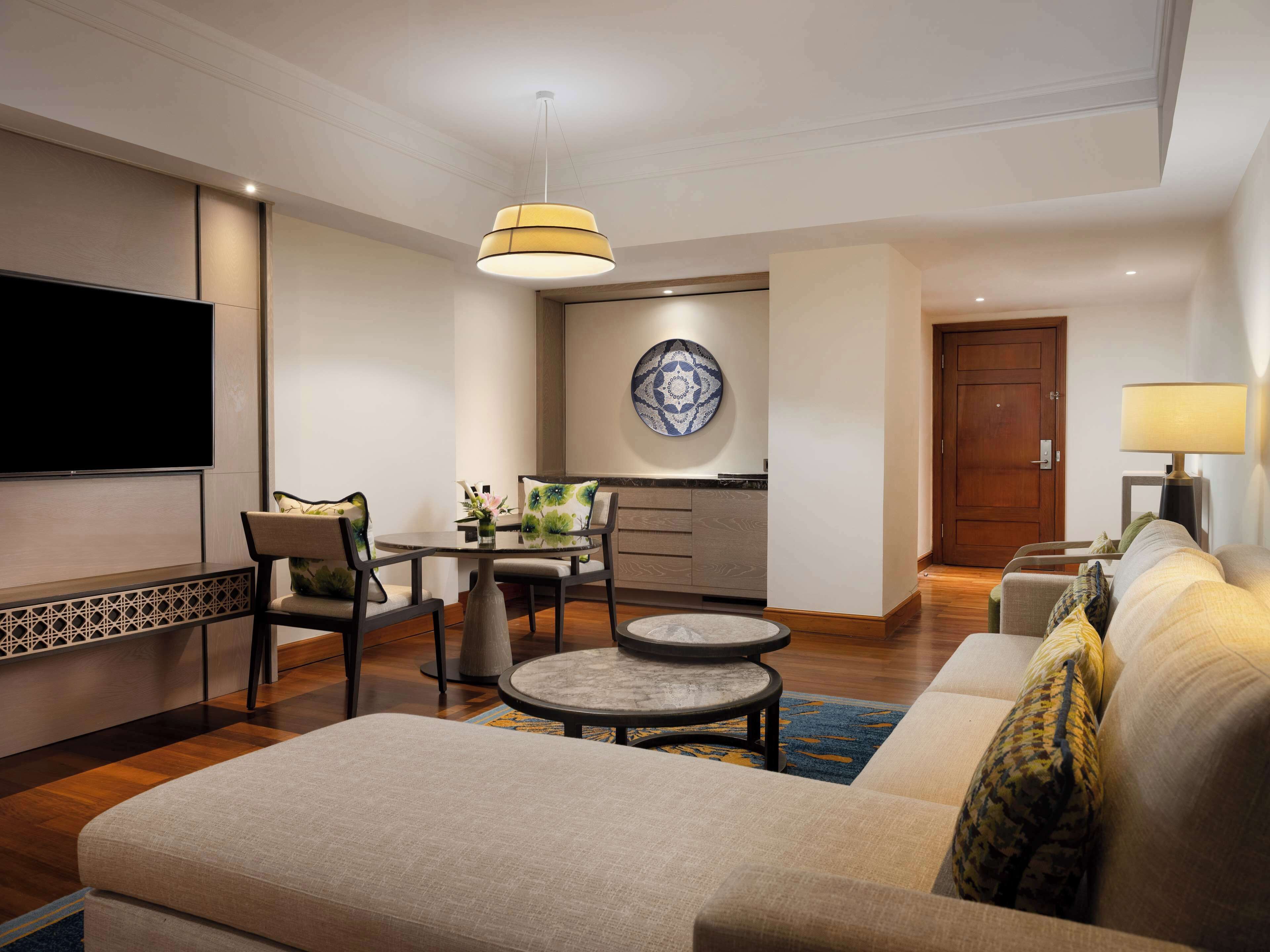 Ein modernes Hotelzimmer mit Flachbildfernseher, einer Couch, einem Esstisch mit Stühlen, einer Wandplatte und einer Lampe.