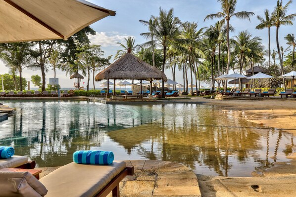 A poolside area with lounge chairs, a thatched-roof structure, and palm trees.