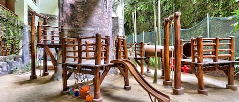 A playground with wooden structures and a sandy floor, surrounded by greenery and a stone wall.