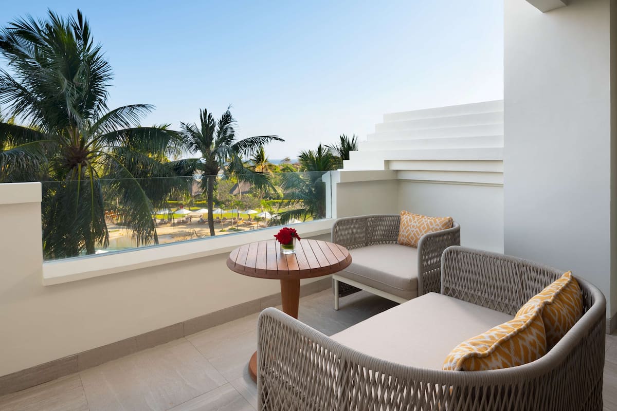 Ein Balkon mit Korbmöbeln, einem kleinen runden Tisch und Blick auf Palmen und einen Strand.