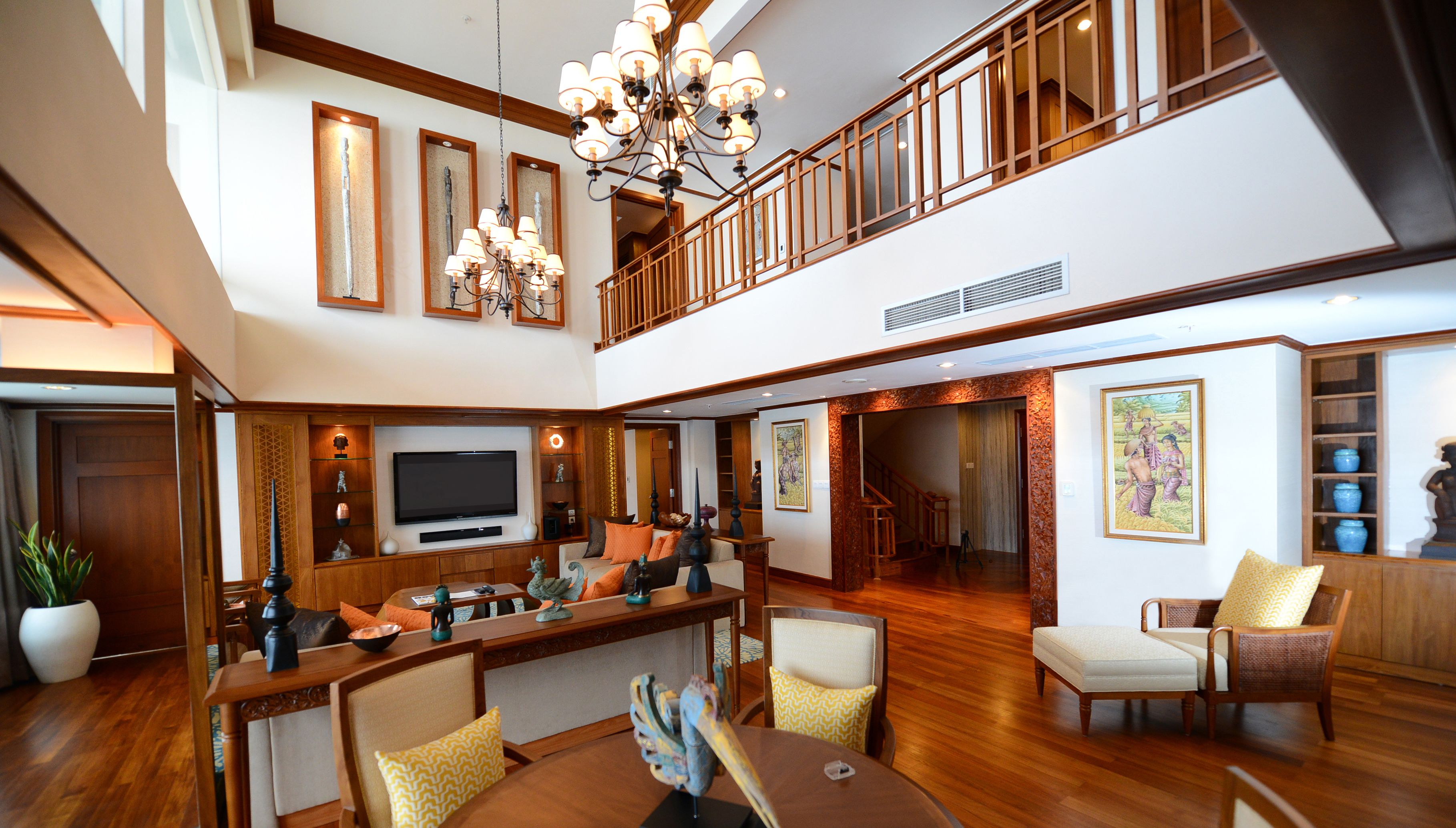 A spacious living area with a high ceiling, a large chandelier, a flat-screen TV, and a wooden staircase leading to an upper level.