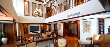 A spacious living area with a high ceiling, a large chandelier, a flat-screen TV, and a wooden staircase leading to an upper level.