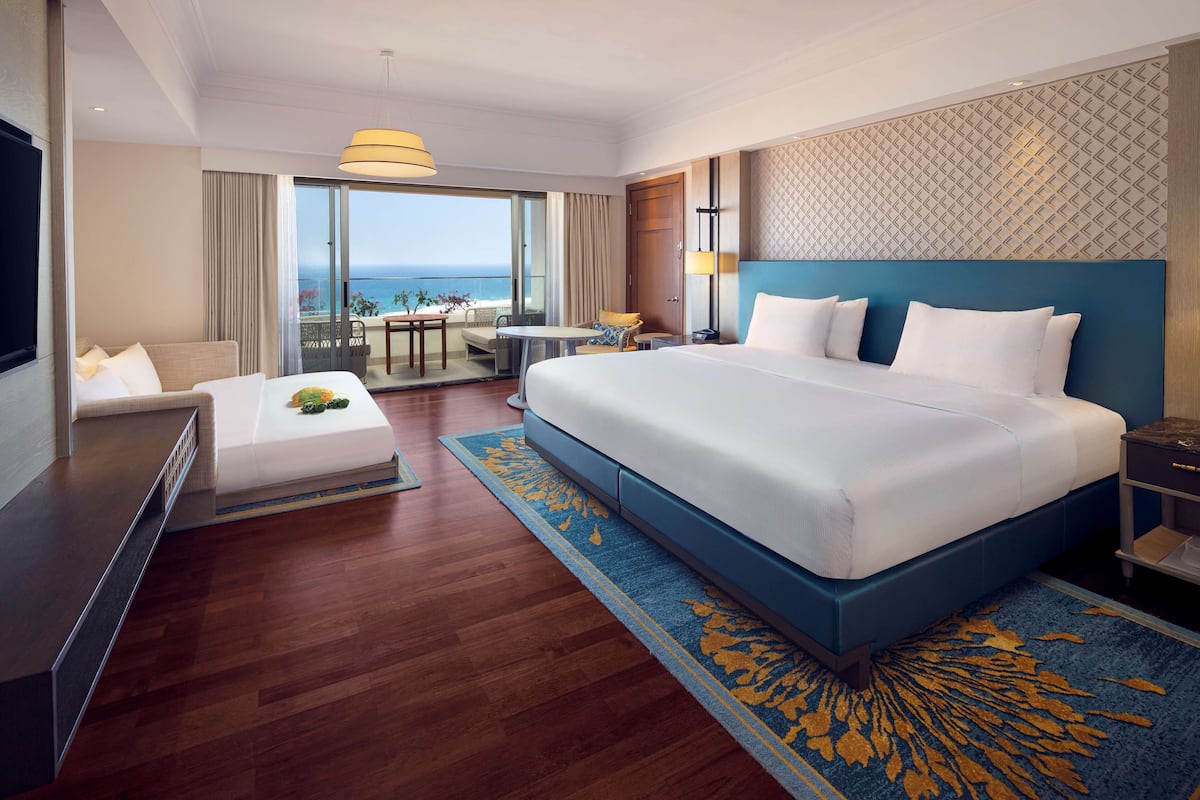 Ein Hotelzimmer mit einem großen Bett, einem Schreibtisch, einem Stuhl, einem Fernseher und einem Balkon mit Meerblick.